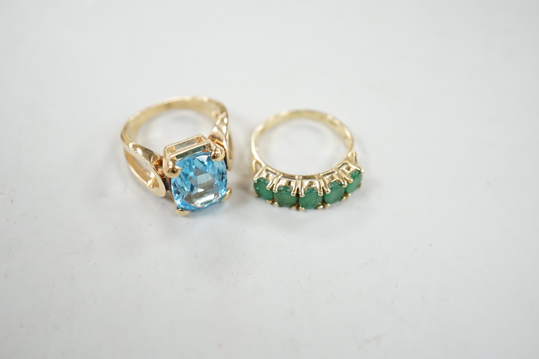 A modern 14k yellow metal and cushion cut blue topaz set dress ring, size K/L and a 14k yellow metal and five stone emerald set half hoop ring, size L/M, gross weight 10.5 grams.